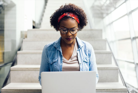 A college-age young woman sitting at the bottom of a staircase working on her computer